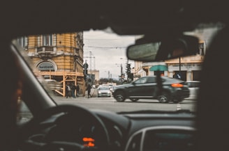 A view from the interior of a car looking out onto a city street, with vehicles and pedestrians traversing an intersection. Buildings with classic architecture and a sign for an agency are visible in the background.