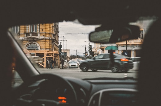 A view from the interior of a car looking out onto a city street, with vehicles and pedestrians traversing an intersection. Buildings with classic architecture and a sign for an agency are visible in the background.