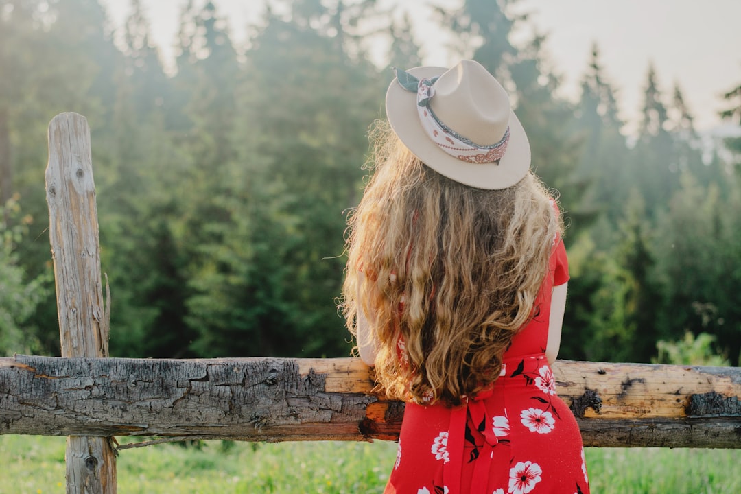 woman in red and white floral dress wearing white cowboy hat standing near brown wooden fence