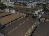 Aerial view of a residential area with several multi-story apartment buildings. The buildings are arranged in rows, featuring brown roofs and beige walls. In the background, taller buildings and industrial structures can be seen, indicating a cityscape. Trees are interspersed among the buildings, and various vehicles are parked in the open areas.
