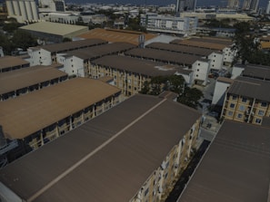 Aerial view of a residential area with several multi-story apartment buildings. The buildings are arranged in rows, featuring brown roofs and beige walls. In the background, taller buildings and industrial structures can be seen, indicating a cityscape. Trees are interspersed among the buildings, and various vehicles are parked in the open areas.