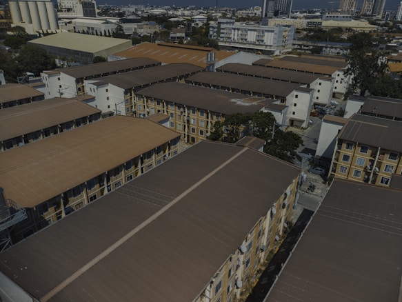 Aerial view of a residential area with several multi-story apartment buildings. The buildings are arranged in rows, featuring brown roofs and beige walls. In the background, taller buildings and industrial structures can be seen, indicating a cityscape. Trees are interspersed among the buildings, and various vehicles are parked in the open areas.