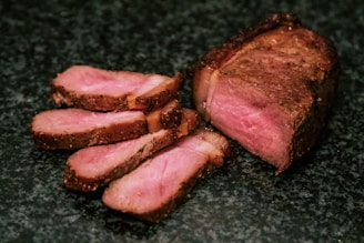 Slices of cooked meat are arranged on a dark surface, showcasing a well-cooked interior with a rich brown crust and a tender pink center.
