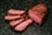 Slices of cooked meat are arranged on a dark surface, showcasing a well-cooked interior with a rich brown crust and a tender pink center.