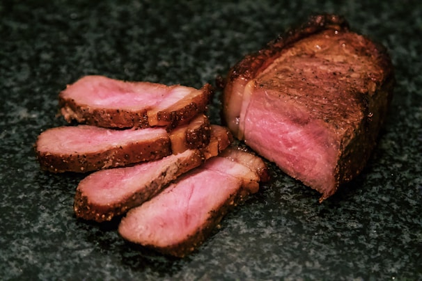 Slices of cooked meat are arranged on a dark surface, showcasing a well-cooked interior with a rich brown crust and a tender pink center.