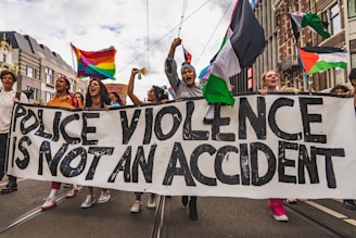 A group of people marching on a street holding a large banner with the text 'Police Violence is Not an Accident'. The marchers are expressing emotions passionately and are carrying flags that include rainbow colors and other designs. The background features urban buildings under a cloudy sky.