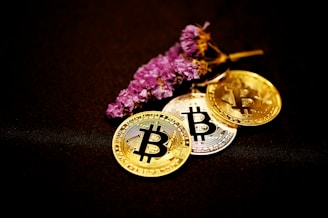 Three metallic Bitcoin coins are arranged on a dark surface alongside a branch of small purple flowers. The coins have intricate circuit-like designs and a prominent Bitcoin symbol in the center. The image combines the digital currency theme with a natural element.