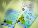 Three books are displayed against a light green background, with two partly facing up and exhibiting colorful covers. The book titled 'Aku Muslim tapi Belum Shalat' has a light blue cover featuring an illustration of a mosque and green hills, accompanied by Islamic-themed text. Another book is titled 'Tata Cara Shalat Menurut Sunnah Nabi' with a white cover and gold and brown text. The third book is shown from the back, featuring a photograph of a man and text describing the content.