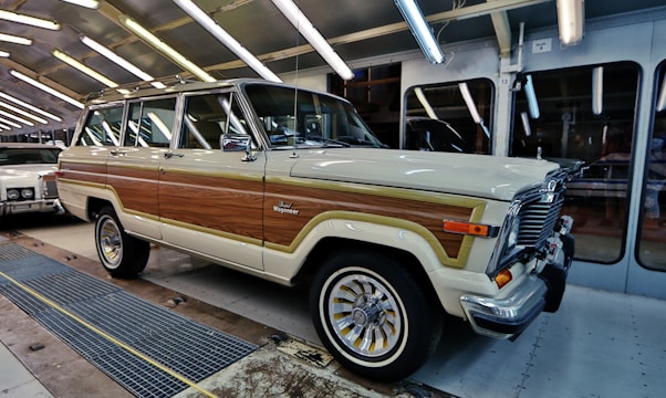 A classic cream and wood-panelled SUV is parked in a well-lit garage or automotive facility. The vehicle features chrome accents and retro-style wheels, with another classic car visible partially in the background.