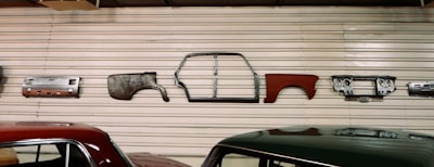 Various vintage car parts are mounted on a corrugated metal wall, including panels, a car frame, and a dashboard. Two cars with polished appearances are parked below the wall.
