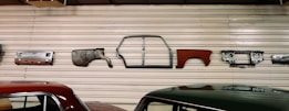 Various vintage car parts are mounted on a corrugated metal wall, including panels, a car frame, and a dashboard. Two cars with polished appearances are parked below the wall.