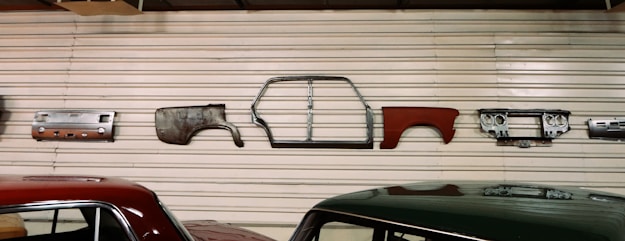 Various vintage car parts are mounted on a corrugated metal wall, including panels, a car frame, and a dashboard. Two cars with polished appearances are parked below the wall.
