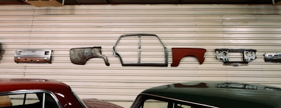 Various vintage car parts are mounted on a corrugated metal wall, including panels, a car frame, and a dashboard. Two cars with polished appearances are parked below the wall.