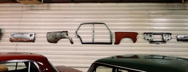 Various vintage car parts are mounted on a corrugated metal wall, including panels, a car frame, and a dashboard. Two cars with polished appearances are parked below the wall.