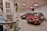 The image depicts a showroom with two red cars displayed prominently. Each car is adorned with red and white balloons. In the foreground, there is an old computer setup on a desk along with some potted plants. Posters and a small plaque are mounted on the walls, and the atmosphere suggests a retro or vintage theme.