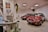 The image depicts a showroom with two red cars displayed prominently. Each car is adorned with red and white balloons. In the foreground, there is an old computer setup on a desk along with some potted plants. Posters and a small plaque are mounted on the walls, and the atmosphere suggests a retro or vintage theme.
