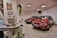 The image depicts a showroom with two red cars displayed prominently. Each car is adorned with red and white balloons. In the foreground, there is an old computer setup on a desk along with some potted plants. Posters and a small plaque are mounted on the walls, and the atmosphere suggests a retro or vintage theme.