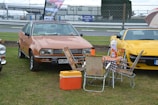 A vintage car is parked on a grassy area alongside a yellow sports car. In front of the vintage car, there is a setup with a foldable table and several chairs that have a floral pattern, along with a thermos and an orange cooler. A checked tablecloth covers the table, and there are barriers and a 'danger prohibited area' sign in the background.