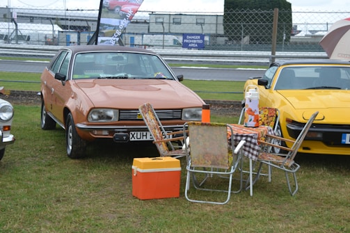 A vintage car is parked on a grassy area alongside a yellow sports car. In front of the vintage car, there is a setup with a foldable table and several chairs that have a floral pattern, along with a thermos and an orange cooler. A checked tablecloth covers the table, and there are barriers and a 'danger prohibited area' sign in the background.