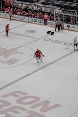 A hockey game is taking place on an ice rink. Players in red and white jerseys are visible, with some players sitting on the bench and others on the ice. Three referees stand near the benches, and the focus is on a goalie dressed in red gear standing alone. Advertisements and logos are printed on the ice surface, and the arena is relatively calm with a few people watching.