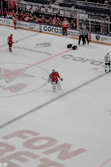 A hockey game is taking place on an ice rink. Players in red and white jerseys are visible, with some players sitting on the bench and others on the ice. Three referees stand near the benches, and the focus is on a goalie dressed in red gear standing alone. Advertisements and logos are printed on the ice surface, and the arena is relatively calm with a few people watching.