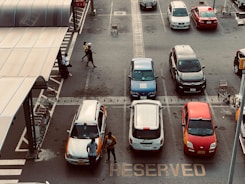 A parking lot with several cars parked in marked spaces. Two people are standing near a car, and another person is walking away from the parking area. Shopping carts are located under a covered area to the side.