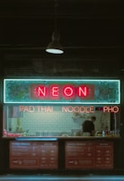 A small restaurant with neon signage displaying 'NEON' above a counter where a person is standing. The signs also advertise 'PAD THAI', 'NOODLE', and 'PHO'. The interior features a backsplash of white subway tiles and various cooking utensils and condiments are visible on the counter.