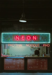 A small restaurant with neon signage displaying 'NEON' above a counter where a person is standing. The signs also advertise 'PAD THAI', 'NOODLE', and 'PHO'. The interior features a backsplash of white subway tiles and various cooking utensils and condiments are visible on the counter.