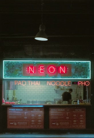 A small restaurant with neon signage displaying 'NEON' above a counter where a person is standing. The signs also advertise 'PAD THAI', 'NOODLE', and 'PHO'. The interior features a backsplash of white subway tiles and various cooking utensils and condiments are visible on the counter.