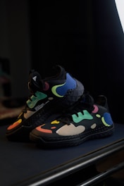 A pair of black athletic shoes with vibrant, multicolored abstract patterns featuring blue, green, yellow, and pink. The shoes are displayed on a dark surface in a dimly lit setting, highlighting their bright, eye-catching design.
