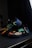 A pair of black athletic shoes with vibrant, multicolored abstract patterns featuring blue, green, yellow, and pink. The shoes are displayed on a dark surface in a dimly lit setting, highlighting their bright, eye-catching design.