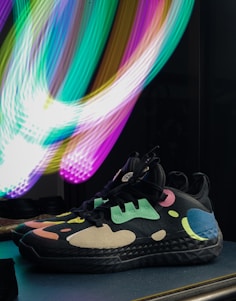 A pair of stylish sneakers with a black base and colorful abstract patterns featuring shades of teal, pink, yellow, and blue. Positioned on a flat surface, the sneakers are highlighted by vibrant, swirling light trails in the background, which create a dynamic contrast.
