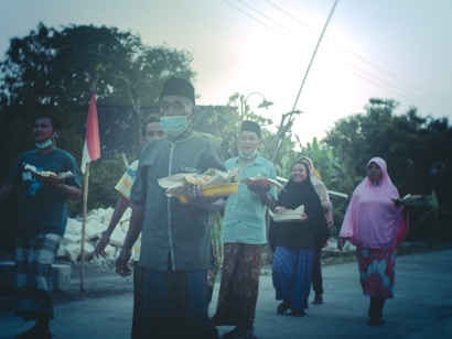 A group of people walking outdoors, each carrying a tray of food. They appear to be in a festive or communal setting, possibly celebrating or participating in a cultural event. The background features greenery and an Indonesian flag. The individuals are wearing a mix of traditional clothing and casual attire.