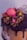 A beautifully decorated chocolate cake with creamy chocolate frosting and colorful heart-shaped candy sprinkles. The cake is topped with a pink macaron and is finished with a glossy chocolate ganache drizzle down the sides.