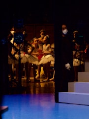 A group of young ballet dancers dressed in white tutus and tiaras are gathered backstage. They appear to be preparing for a performance. The lighting is dim, with subtle stage highlights illuminating the dancers. An individual wearing a dark outfit and a face mask stands to one side, possibly a stagehand or instructor.
