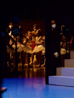 A group of young ballet dancers dressed in white tutus and tiaras are gathered backstage. They appear to be preparing for a performance. The lighting is dim, with subtle stage highlights illuminating the dancers. An individual wearing a dark outfit and a face mask stands to one side, possibly a stagehand or instructor.
