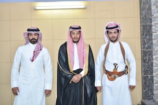 Three men stand side by side in traditional Middle Eastern attire. The man in the middle wears a black robe with gold trim over a white garment, while the other two men wear white thobes. The men on the left and right have traditional red and white checkered headscarves, and the man on the right also has a gold and brown belt with a ceremonial dagger.