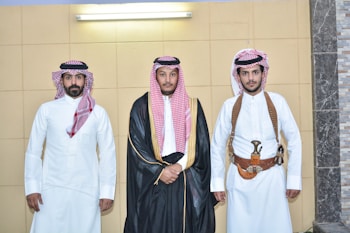 Three men stand side by side in traditional Middle Eastern attire. The man in the middle wears a black robe with gold trim over a white garment, while the other two men wear white thobes. The men on the left and right have traditional red and white checkered headscarves, and the man on the right also has a gold and brown belt with a ceremonial dagger.