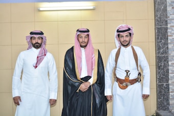 Three men stand side by side in traditional Middle Eastern attire. The man in the middle wears a black robe with gold trim over a white garment, while the other two men wear white thobes. The men on the left and right have traditional red and white checkered headscarves, and the man on the right also has a gold and brown belt with a ceremonial dagger.