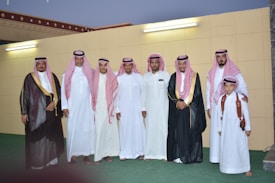 A group of men and a boy dressed in traditional Middle Eastern attire pose together in front of a wall with two light fixtures. The adults wear white thobes and red and white checkered keffiyehs, and some wear black bishts with gold trim. One man is holding a young boy wearing a white thobe and red checkered keffiyeh, with a brown leather shoulder strap.