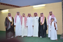 A group of men and a boy dressed in traditional Middle Eastern attire pose together in front of a wall with two light fixtures. The adults wear white thobes and red and white checkered keffiyehs, and some wear black bishts with gold trim. One man is holding a young boy wearing a white thobe and red checkered keffiyeh, with a brown leather shoulder strap.