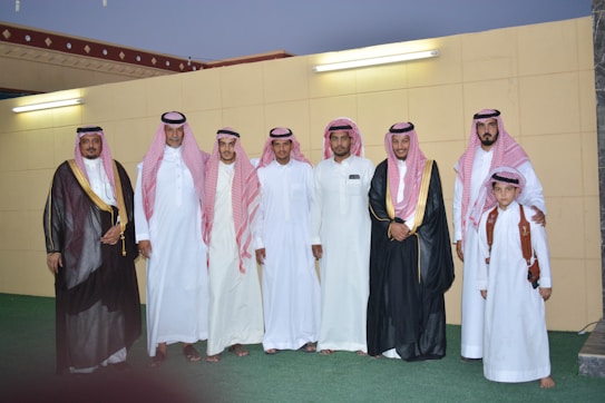A group of men and a boy dressed in traditional Middle Eastern attire pose together in front of a wall with two light fixtures. The adults wear white thobes and red and white checkered keffiyehs, and some wear black bishts with gold trim. One man is holding a young boy wearing a white thobe and red checkered keffiyeh, with a brown leather shoulder strap.