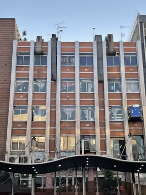 A multi-story building with a facade featuring rectangular windows aligned in rows and columns. The exterior is composed of brown panels with metallic vertical elements. Rooftop antennas and ventilation pipes are visible, and there is a canopy structure at the building's entrance.