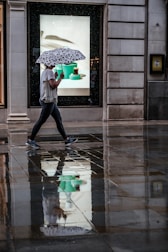 A person is walking on a wet sidewalk, holding an umbrella with a black-and-white pattern. The ground reflects their image along with the surrounding buildings and a brightly lit shop window advertising some items. The scene suggests recent rain and the person's casual attire indicates a relaxed setting.