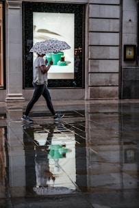 A person is walking on a wet sidewalk, holding an umbrella with a black-and-white pattern. The ground reflects their image along with the surrounding buildings and a brightly lit shop window advertising some items. The scene suggests recent rain and the person's casual attire indicates a relaxed setting.