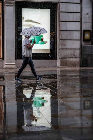 A person is walking on a wet sidewalk, holding an umbrella with a black-and-white pattern. The ground reflects their image along with the surrounding buildings and a brightly lit shop window advertising some items. The scene suggests recent rain and the person's casual attire indicates a relaxed setting.
