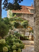 A charming garden courtyard features stone walls and a tiled roof, with lush green foliage and potted plants. The setting has a rustic feel, enhanced by the presence of a decorative gnome statue on a barrel and an ornate bird sculpture. Sunlight filters through the trees, casting dappled shadows on the stone pathway.