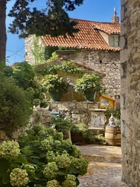 A charming garden courtyard features stone walls and a tiled roof, with lush green foliage and potted plants. The setting has a rustic feel, enhanced by the presence of a decorative gnome statue on a barrel and an ornate bird sculpture. Sunlight filters through the trees, casting dappled shadows on the stone pathway.