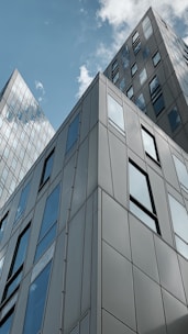Tall modern buildings with reflective glass windows reach up to a partly cloudy sky. The architecture features clean lines and angular designs, showcasing urban development and contemporary style.