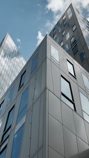 Tall modern buildings with reflective glass windows reach up to a partly cloudy sky. The architecture features clean lines and angular designs, showcasing urban development and contemporary style.